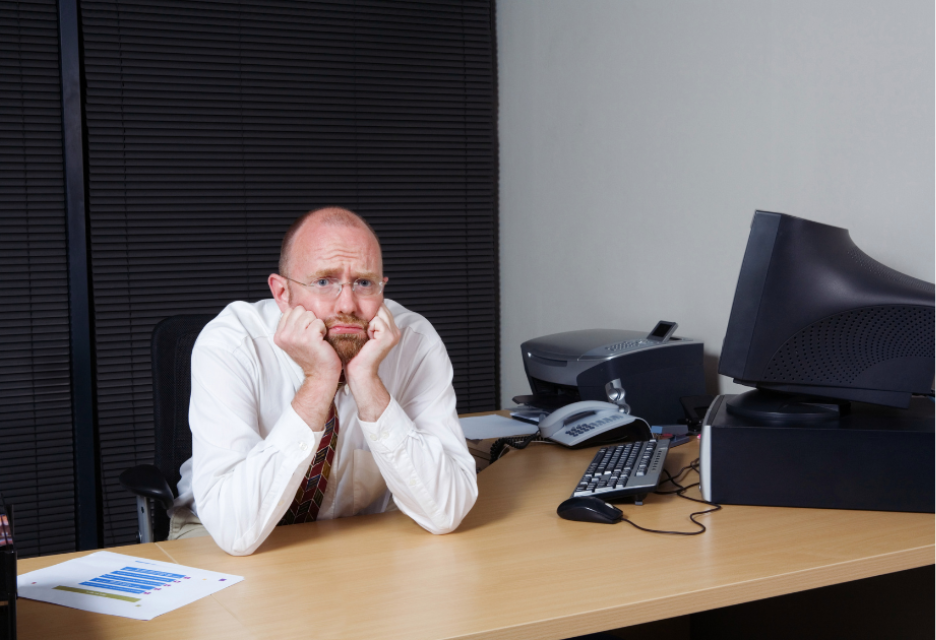 a man sits with his elbows on his computer desk with his chin in his hand. He has a pouty look on his face